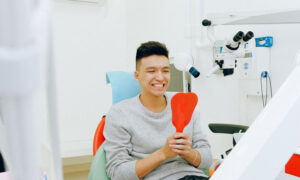 A man sits in a dentist chair, happy with the results of his latest check up, as holds a mirror inspecting his teeth.