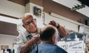 A man nearing the age of retirement is working as a barber, cutting the sideburns of another man in his barber chair.