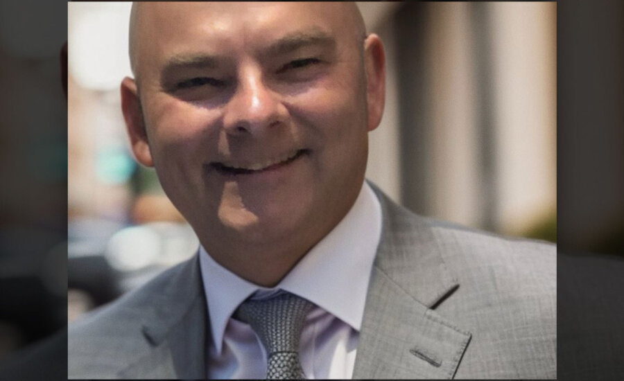 Financial Planner Kenneth Doll smiles into a camera with a blurred city background in the image.