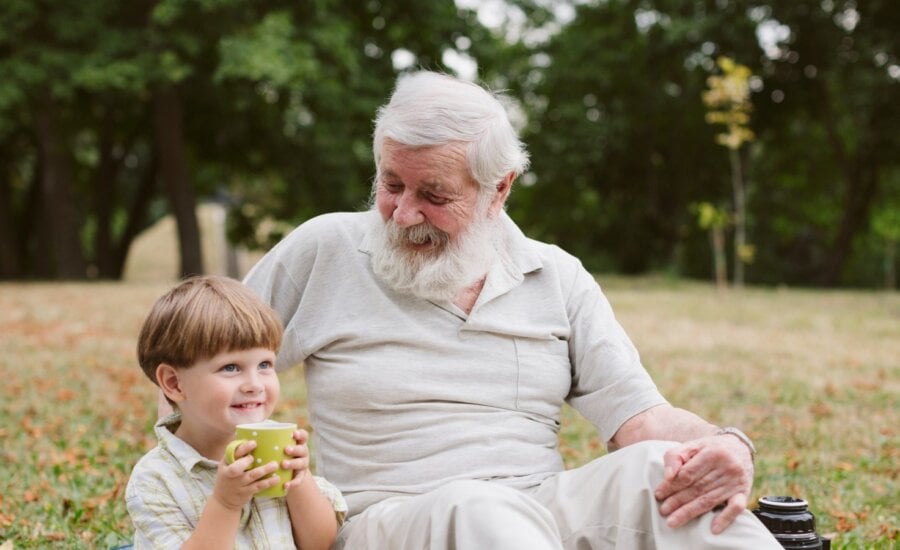 A man sits in the grass with this grandson, who is drinking from a cup