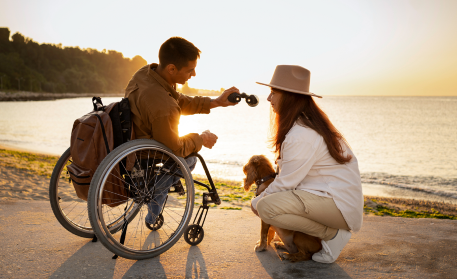 A young man in a wheelchair on a beach with a woman and small dog