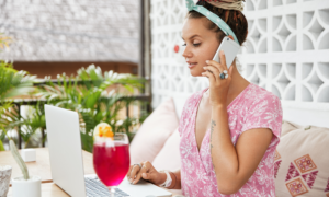 A woman uses her phone and laptop on a sunny patio with a cocktail