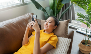 A young woman smiles at her phone while lying on a sofa