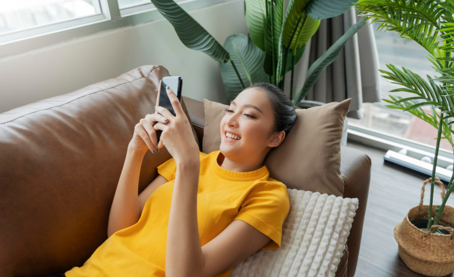 A young woman smiles at her phone while lying on a sofa