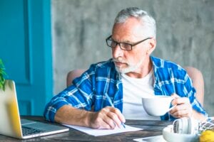 senior-man-looking-laptop-holding-pen-making-notes