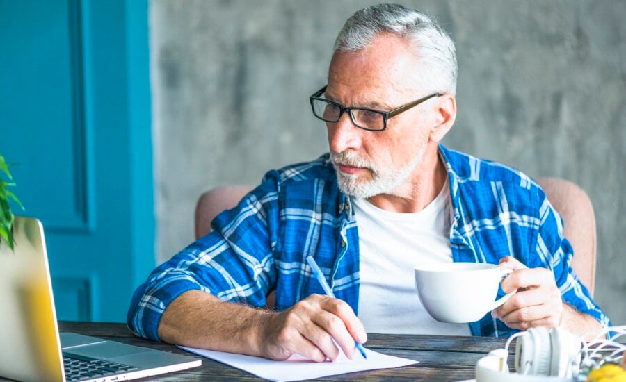 senior-man-looking-laptop-holding-pen-making-notes