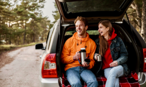 A young man and woman sit in the open trunk of their hatchback on a rural road